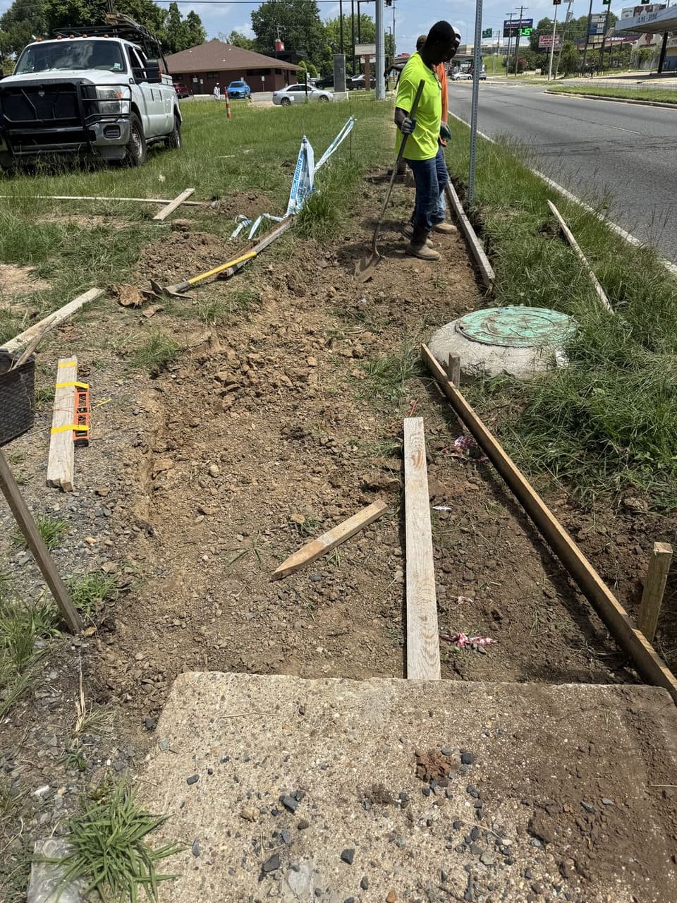 Silverback crew grading subgrade between set wood forms for a new sidewalk along a highway corridor, working around an existing utility structure